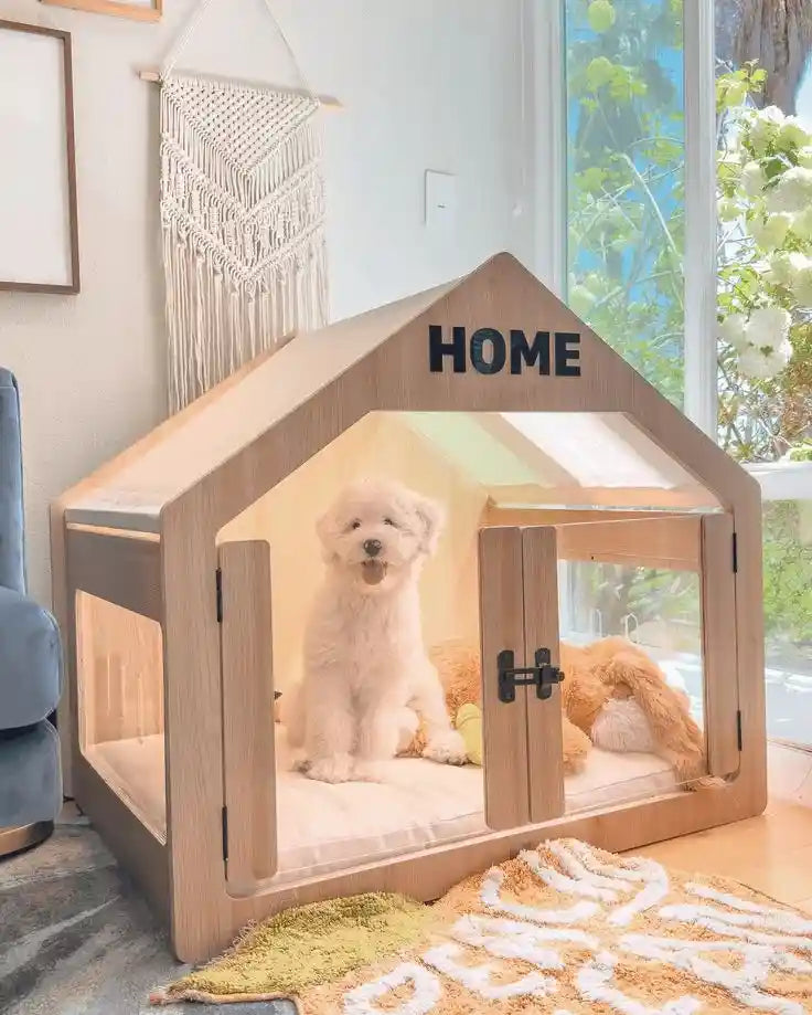 A fluffy white dog sitting happily inside a modern, house-shaped wooden indoor dog crate with clear acrylic panels in a bright, tranquil living room.